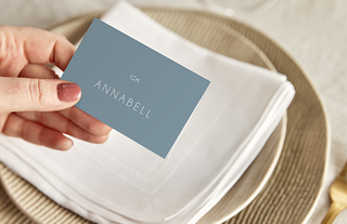 Tischkarte mit christlichem Fisch Symbol und Text "Annabell" wird von einer Hand gehalten, ein Tischgedeck im Hintergrund