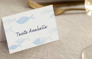Nahaufnahme einer Tischkarte mit Fisch-Symbolen designt, mit Text "Tante Annabell", aufgestellt auf einem Tisch